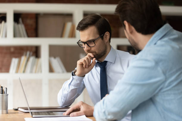 Thoughtful young male boss leader in eyewear looking at laptop screen, discussing project problem solution with colleague at brainstorming meeting. Two managers developing growth strategy in office.